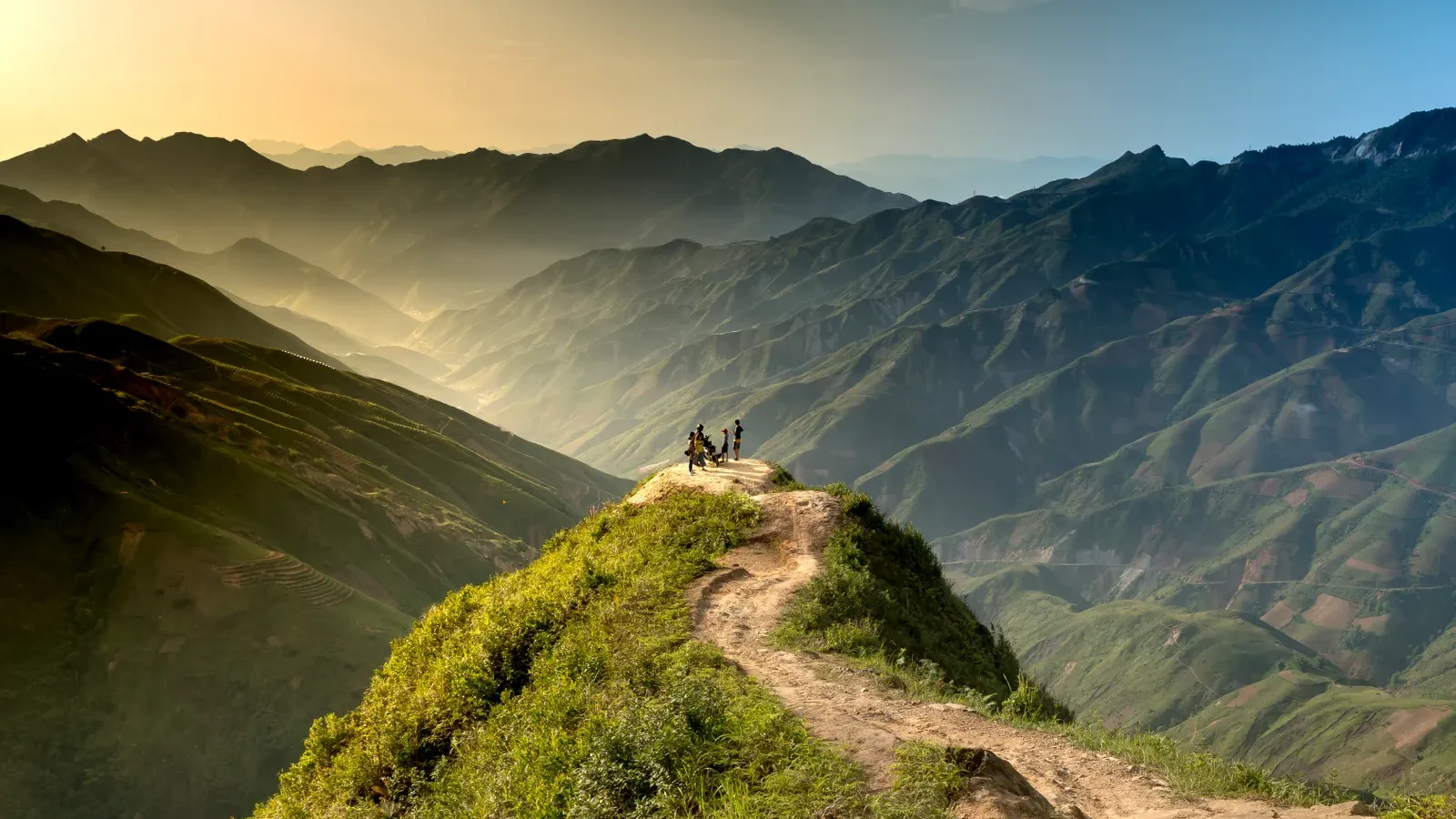 Hikers on a ridge trail overlooking layered mountains at sunrise