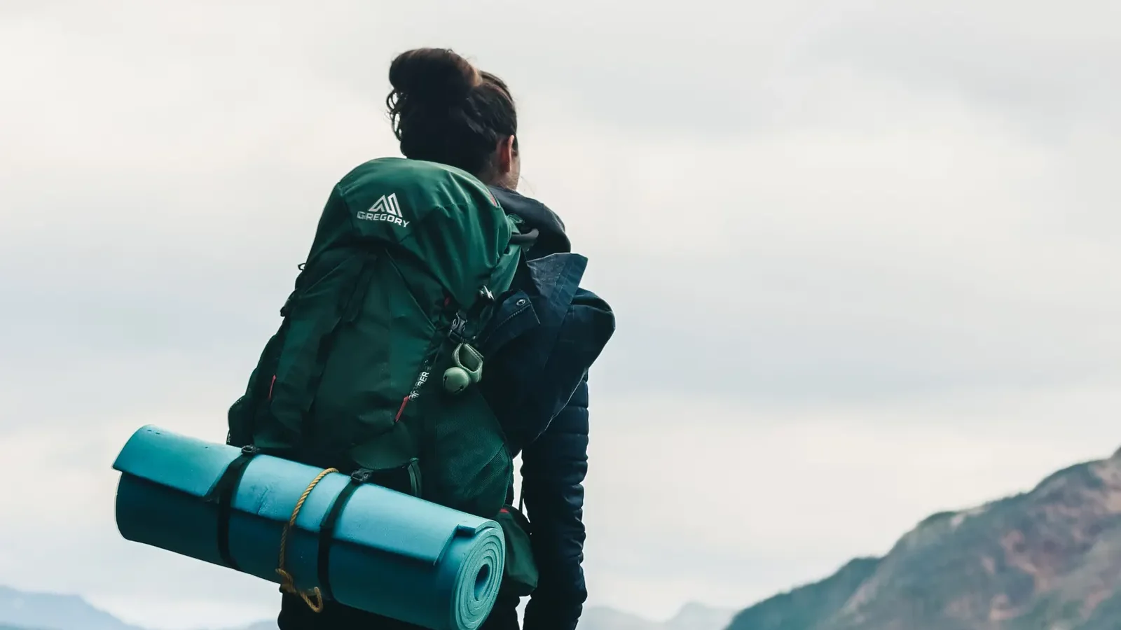 Hiker carrying a large backpack with a rolled sleeping pad, looking at the mountains