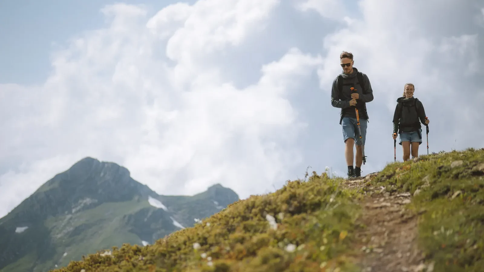 Two hikers using trekking poles on a grassy mountain ridge under big clouds