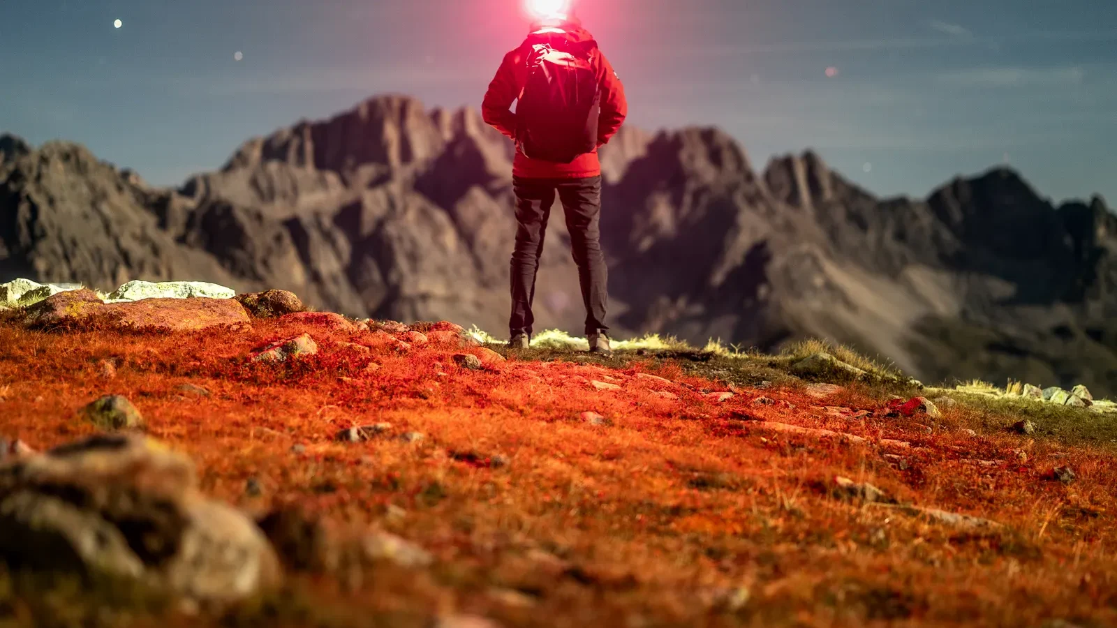 Hiker with a lit headlamp facing a mountain landscape at dusk