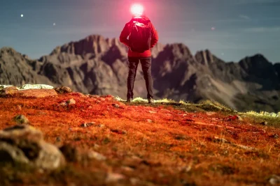 Hiker with a lit headlamp facing a mountain landscape at dusk