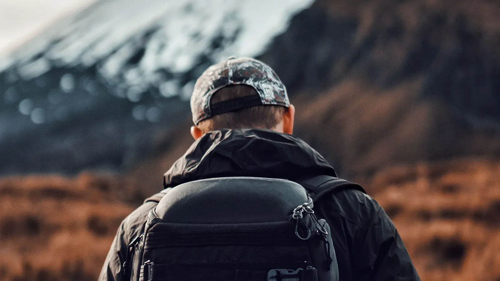 Back view of a hiker wearing a black backpack facing a snow-capped mountain