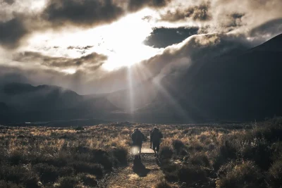 Two hikers crossing a high valley under dramatic storm clouds and low sunlight