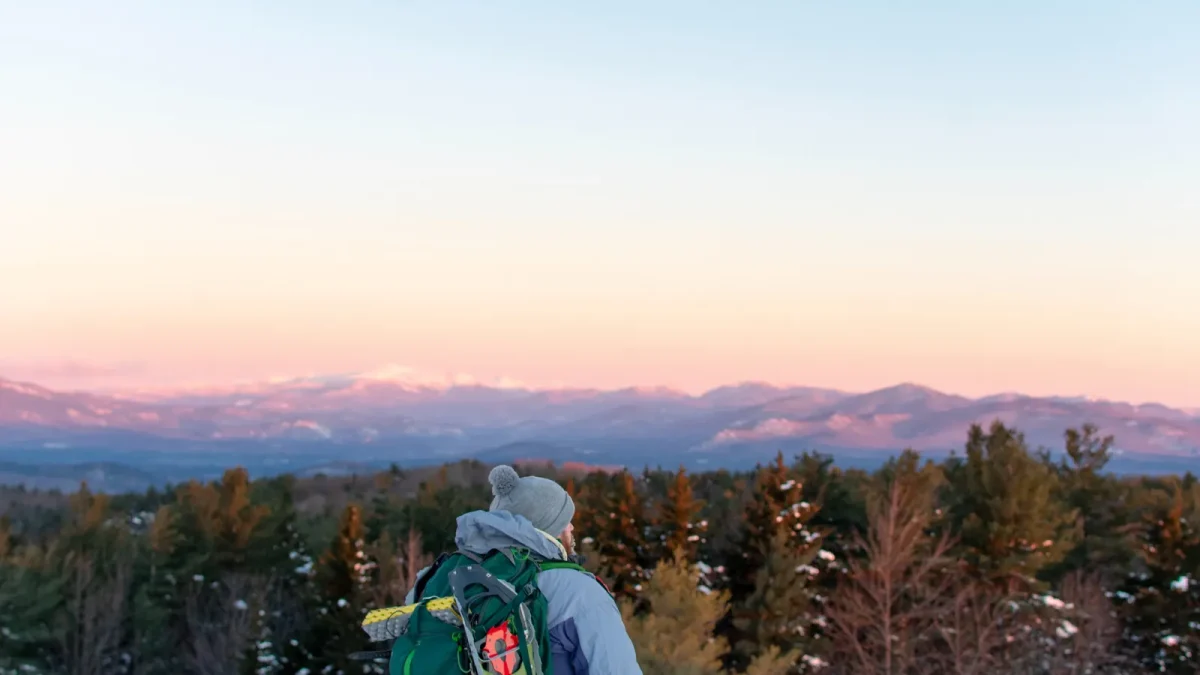 Backpacker with a green pack watching the sun set over snow-dusted mountains in New Hampshire