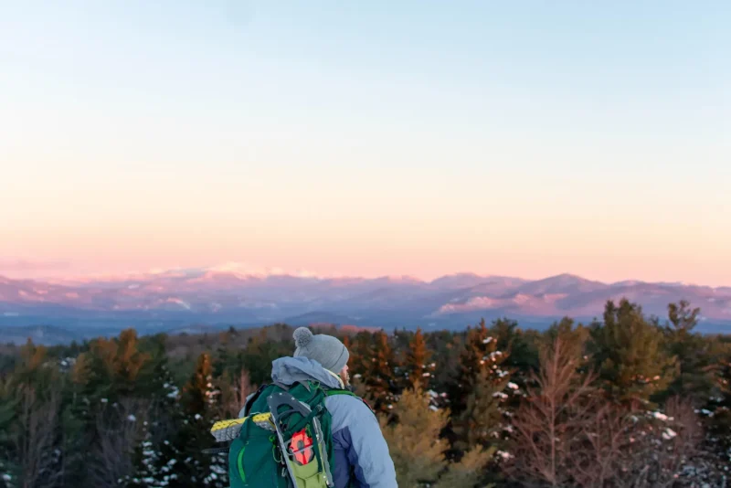 Backpacker with a green pack watching the sun set over snow-dusted mountains in New Hampshire