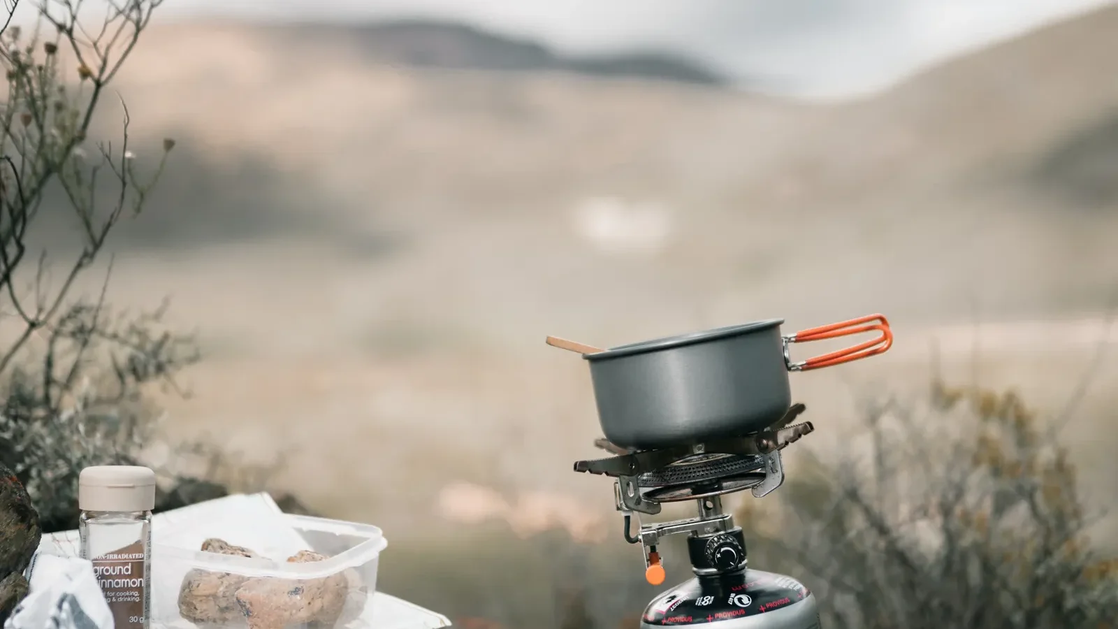 Small camp stove and pot set up on rocky ground with a misty mountain valley behind it