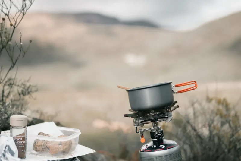 Small camp stove and pot set up on rocky ground with a misty mountain valley behind it