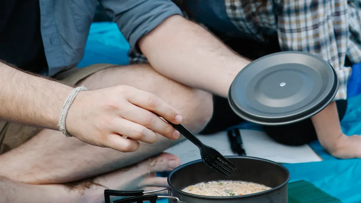 Two campers sharing a pot with its lid set aside on a camping mat