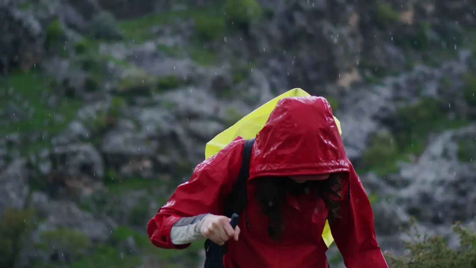 Hiker in a red hooded jacket climbing a rain-soaked, rocky mountain trail