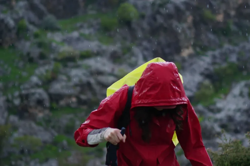 Hiker in a red hooded jacket climbing a rain-soaked, rocky mountain trail