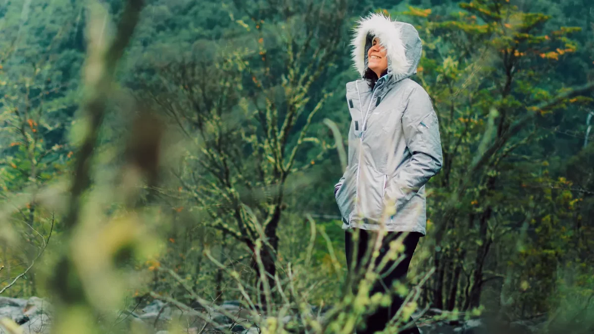 Hiker in a light gray hooded jacket looking up amid green forest trees