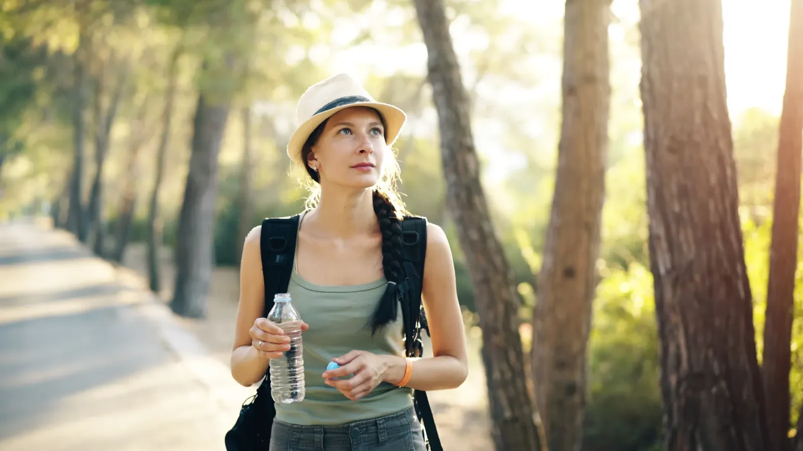Woman in a hat and olive tank top drinking water on a sunlit forest trail
