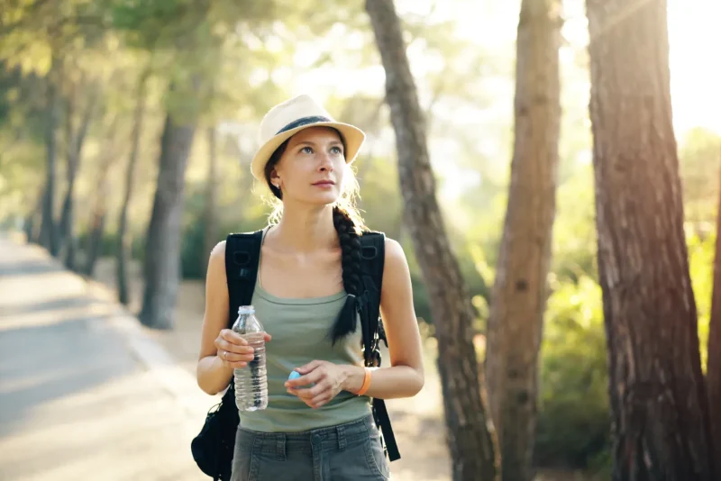 Woman in a hat and olive tank top drinking water on a sunlit forest trail