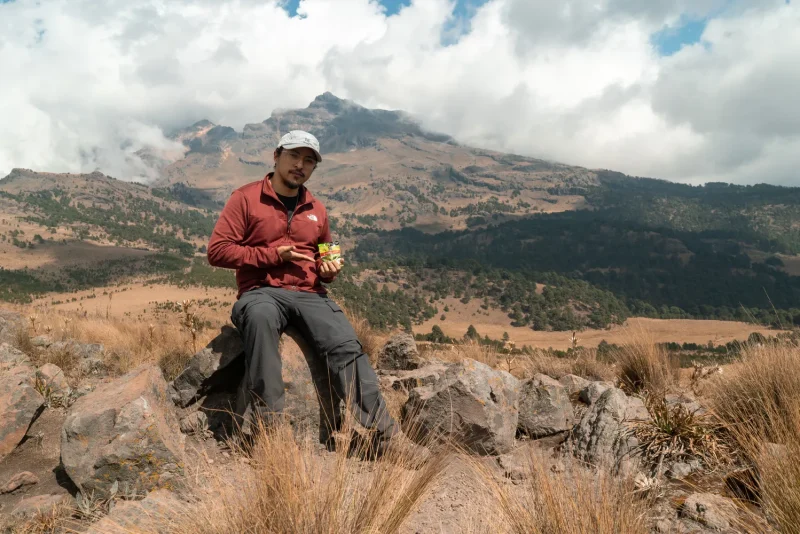 Man sitting on rocks eating a snack with a wide mountain view behind him