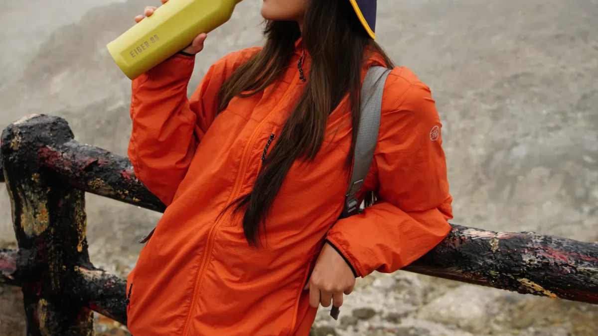 Woman in an orange jacket drinking water from a bottle on a misty mountain trail