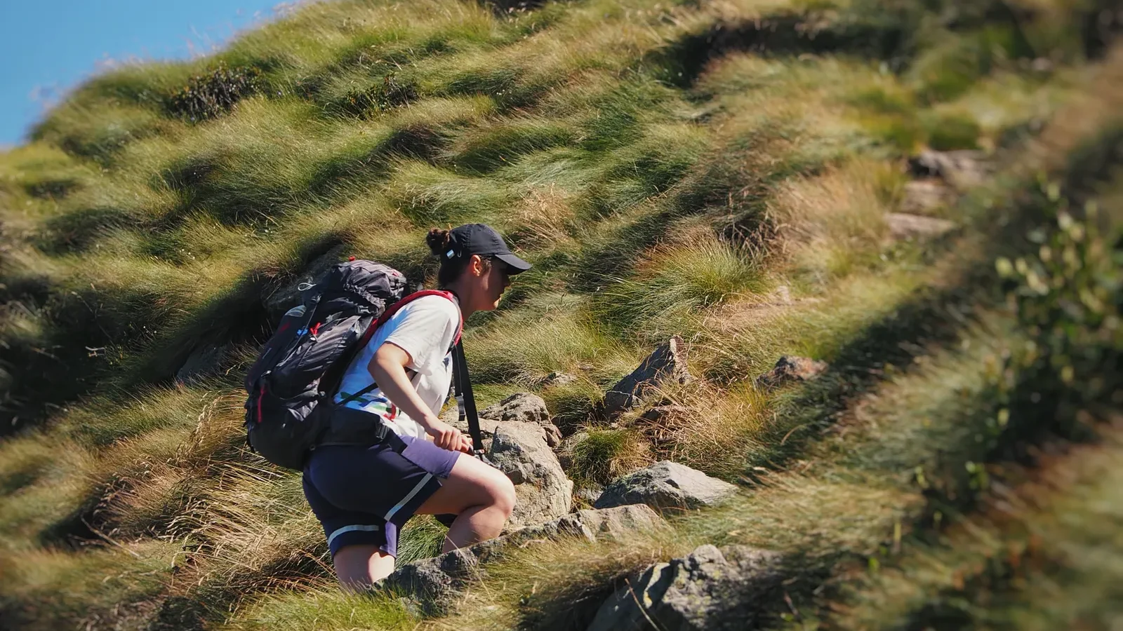 Woman with a backpack climbing a steep, grassy and rocky mountain trail