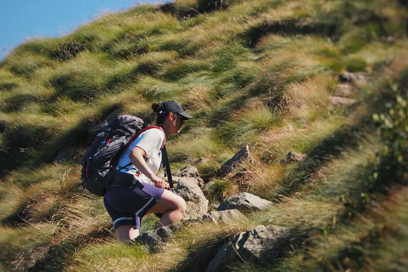 Woman with a backpack climbing a steep, grassy and rocky mountain trail